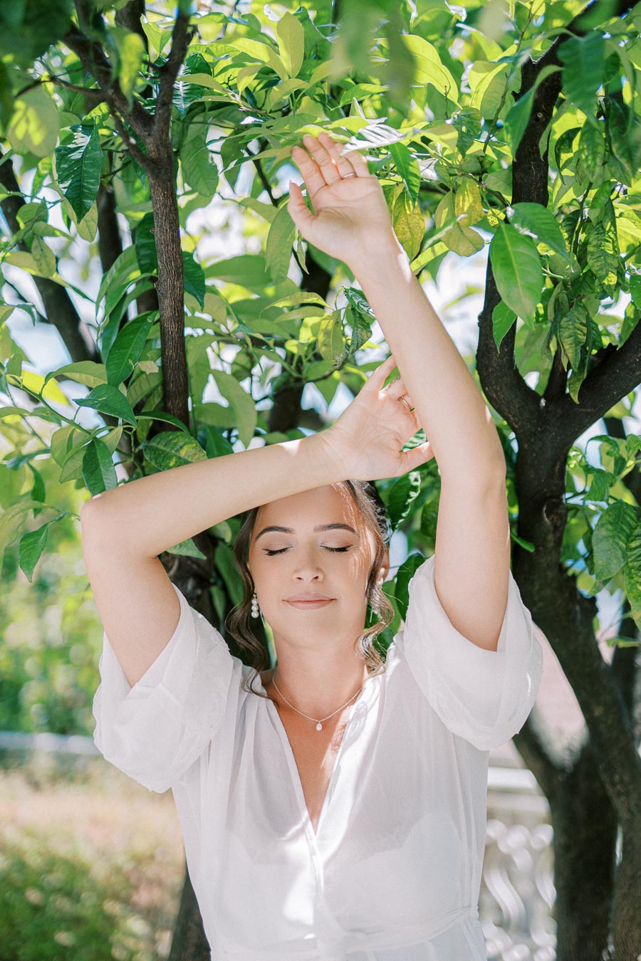 A woman wearing a white dress stands with her eyes closed, raising her arms among lush green leaves and branches in a sunlit outdoor setting.