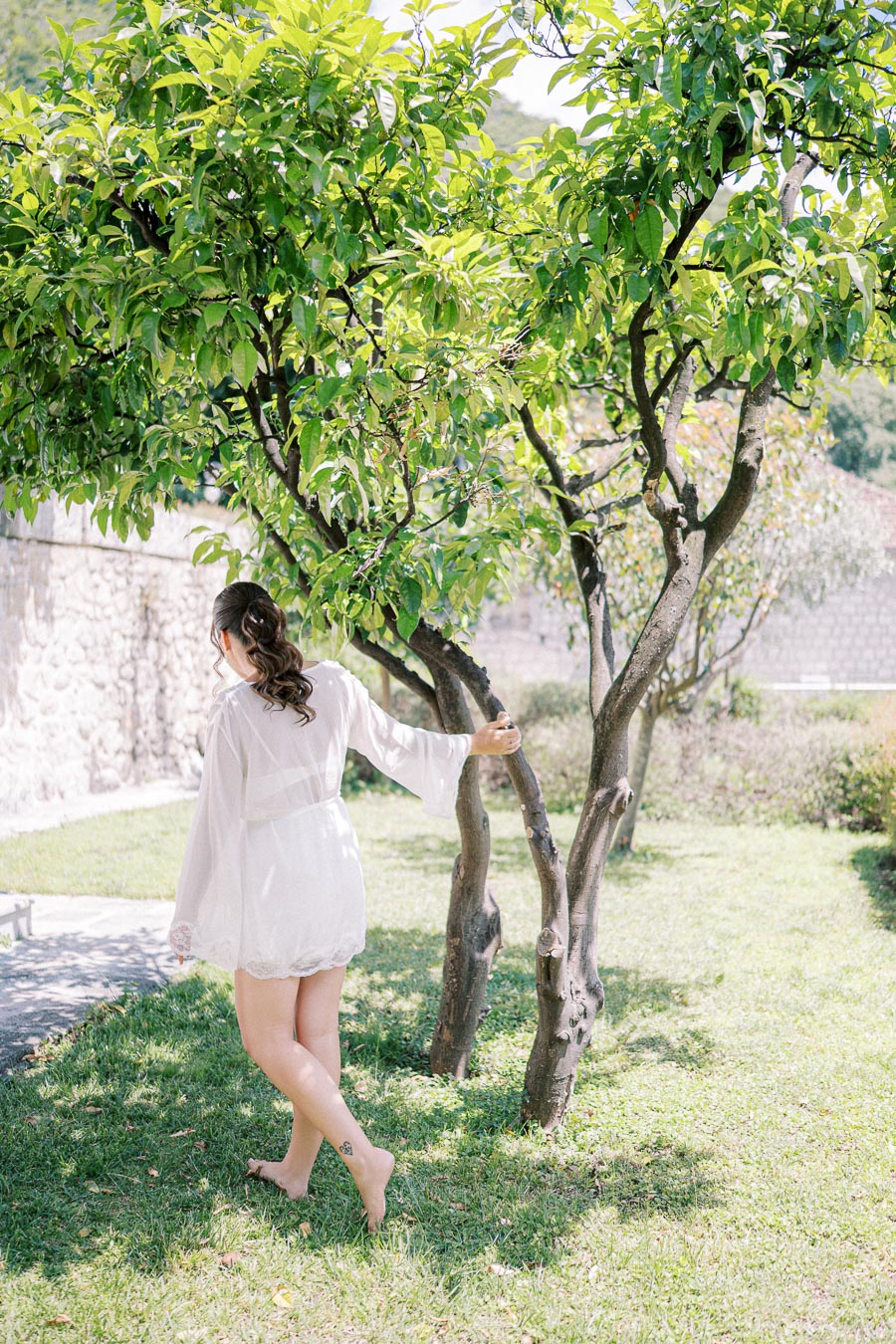 A woman in a white robe gracefully walks barefoot on a lush green lawn, holding onto a tree branch in a serene garden setting.