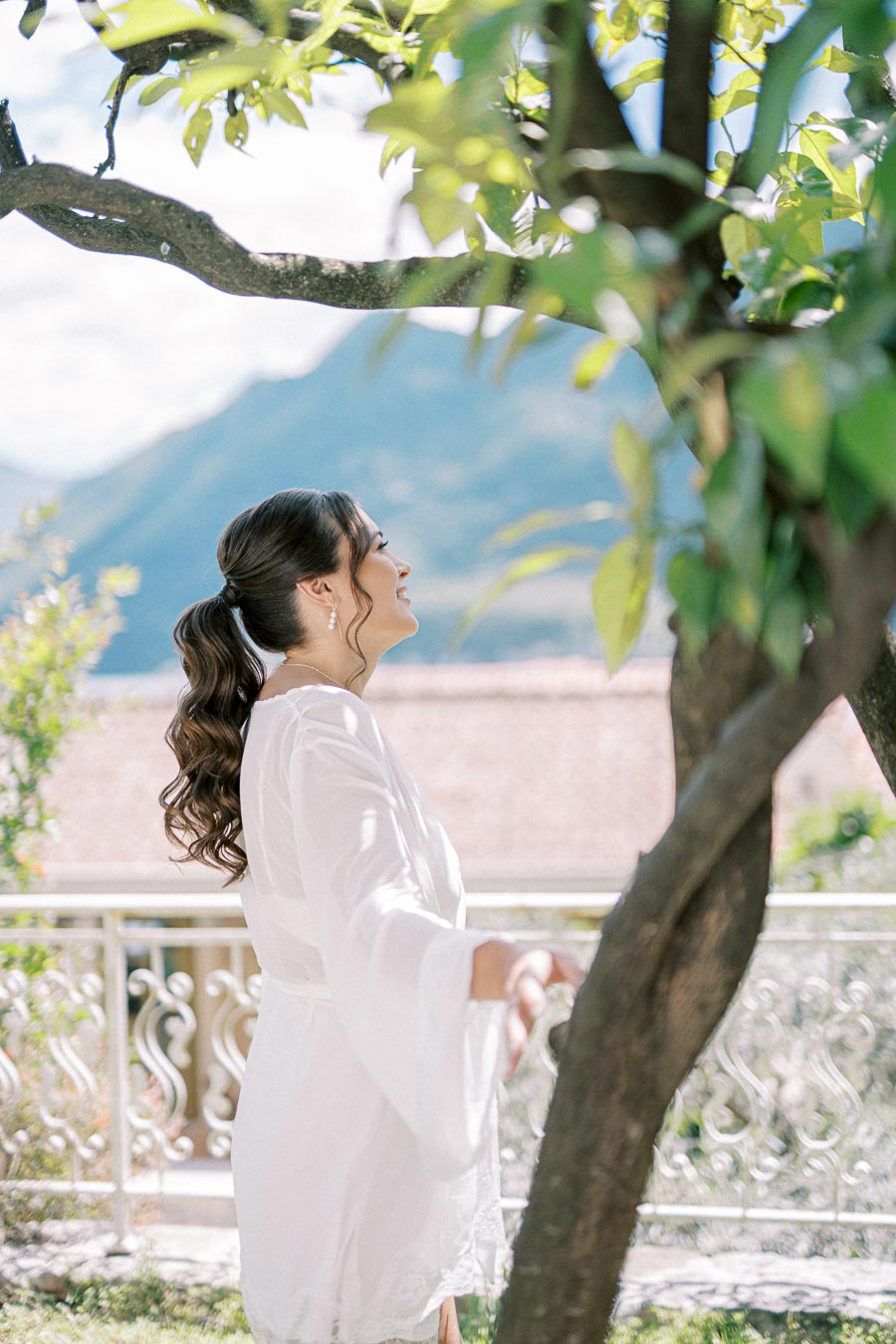 A woman in a white robe smiling joyfully under a tree with mountains in the background.