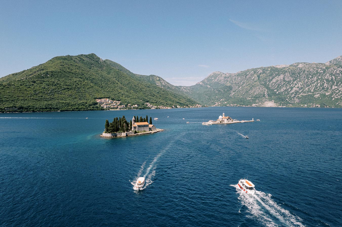 Coastal scenery with two small islands in a bay, featuring historic buildings surrounded by lush greenery and blue waters, boats navigating around the islands, and mountainous landscape in the background under a clear sky.