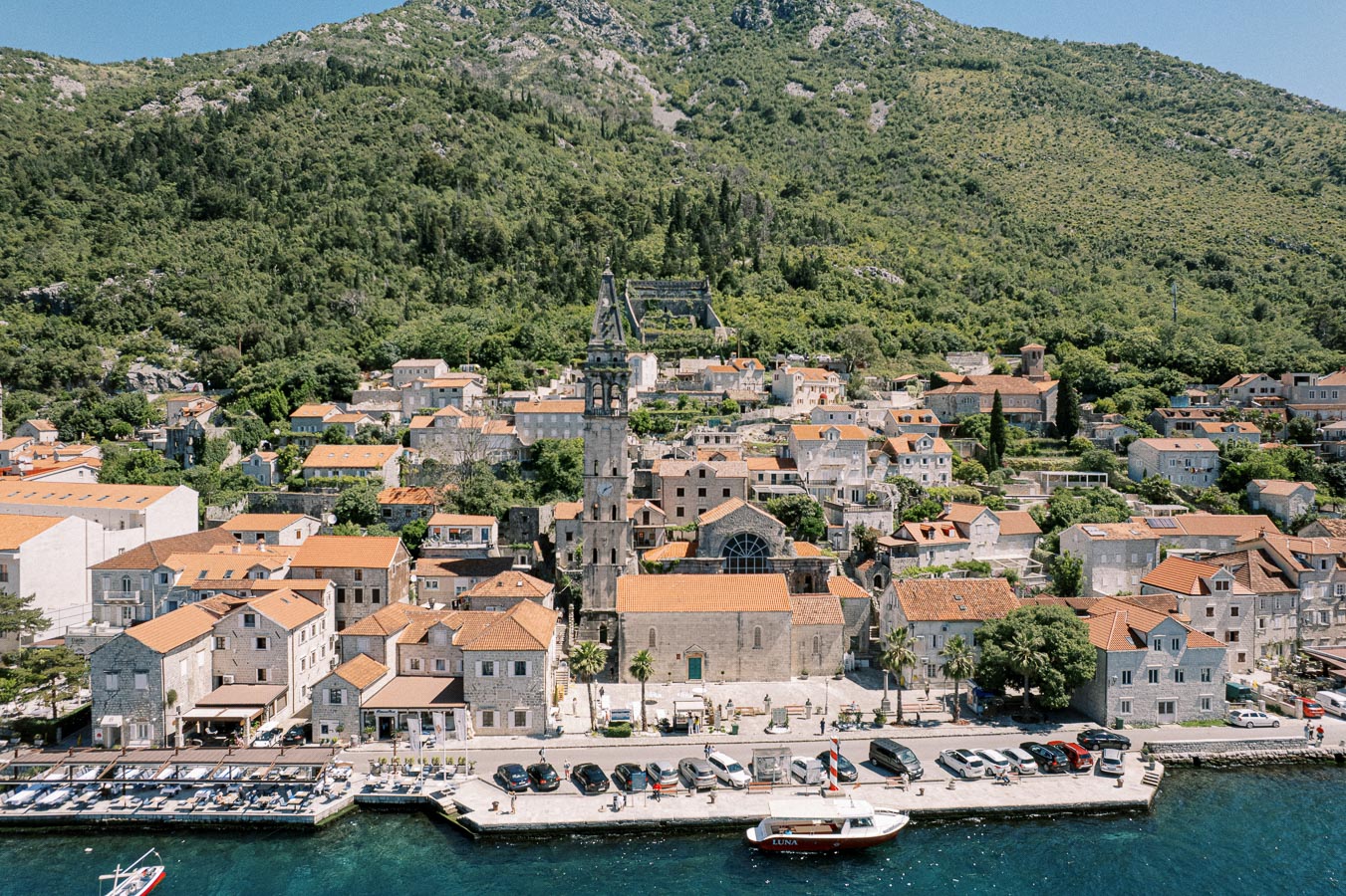 Aerial view of a picturesque Mediterranean coastal town with stone buildings and orange-tiled roofs, nestled at the base of a lush green hillside, showcasing a historic church with a prominent bell tower by the waterfront, lined with parked cars and small boats on a sunny day.
