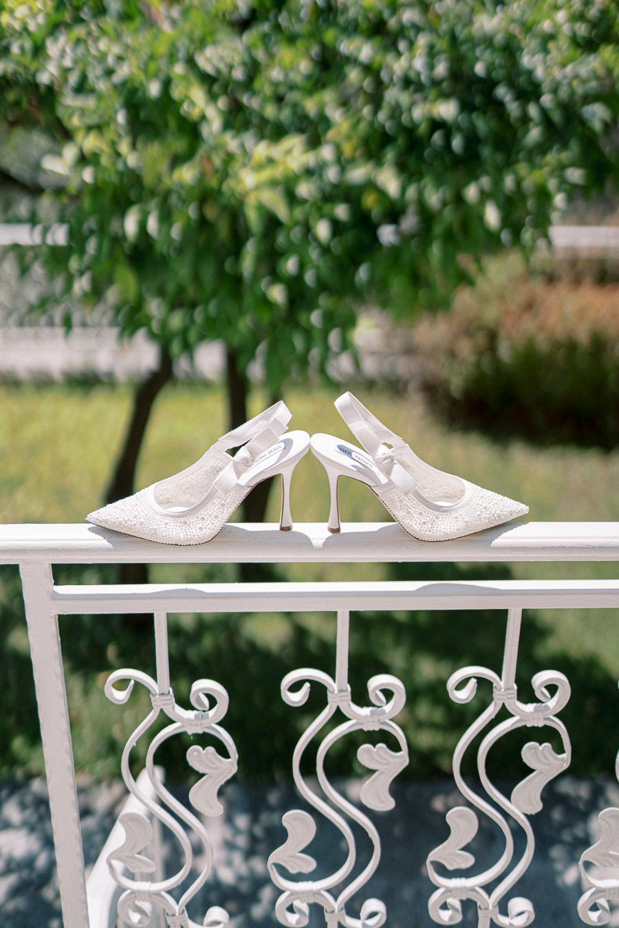 Elegant white high-heeled bridal shoes with intricate lace design resting on a decorative white railing, with a lush green garden background.