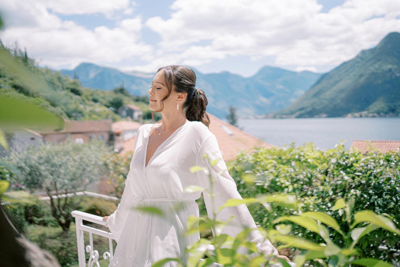 A woman in a white robe enjoying a serene garden with lush greenery, set against a backdrop of picturesque mountains and a tranquil lake under a partly cloudy sky.