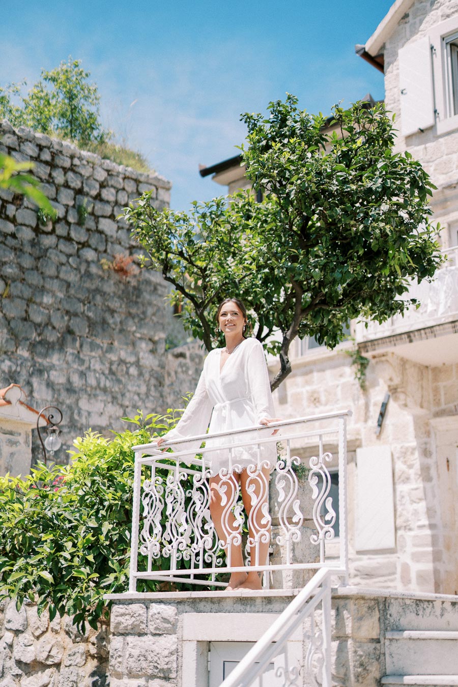 Young woman in a white dress standing on a charming stone balcony surrounded by greenery, with a backdrop of rustic stone buildings under a clear blue sky.