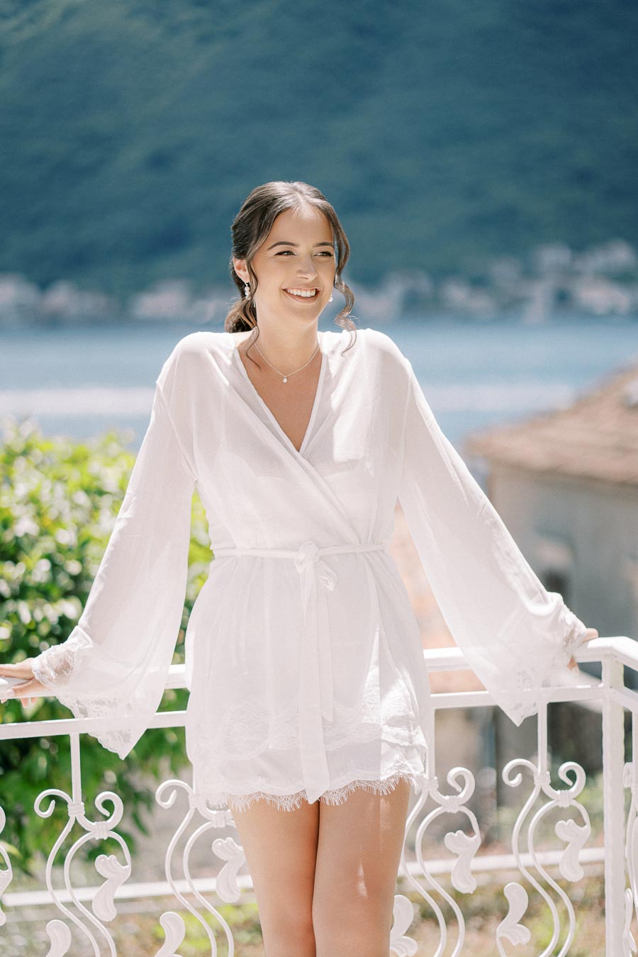 Smiling woman in a white robe standing on a balcony with a scenic mountain and lake view in the background.