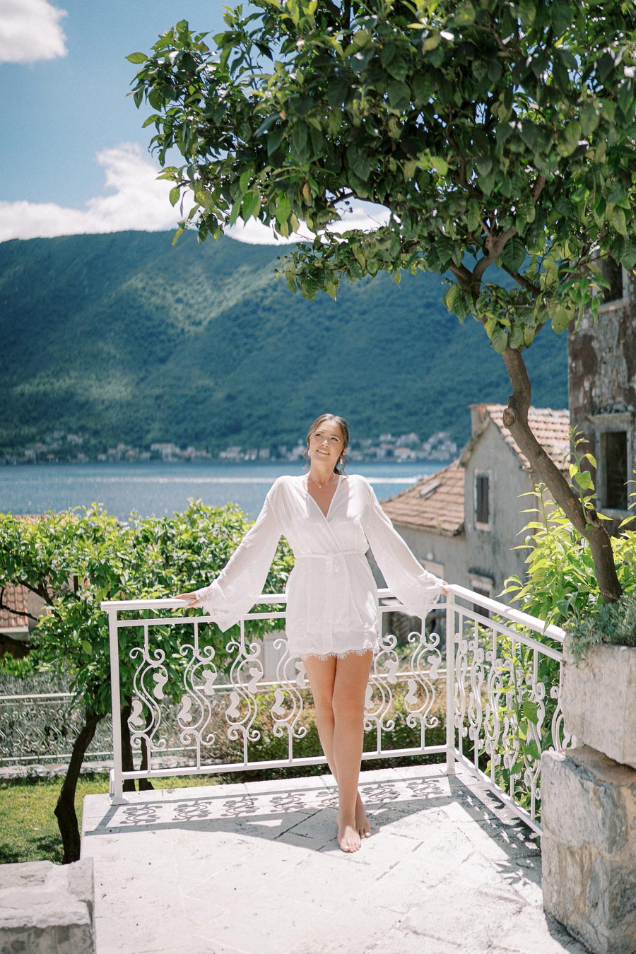 A woman in a white dress stands on a stone balcony overlooking a scenic view of a lush mountain landscape and a calm body of water, with bright green foliage and a clear blue sky.