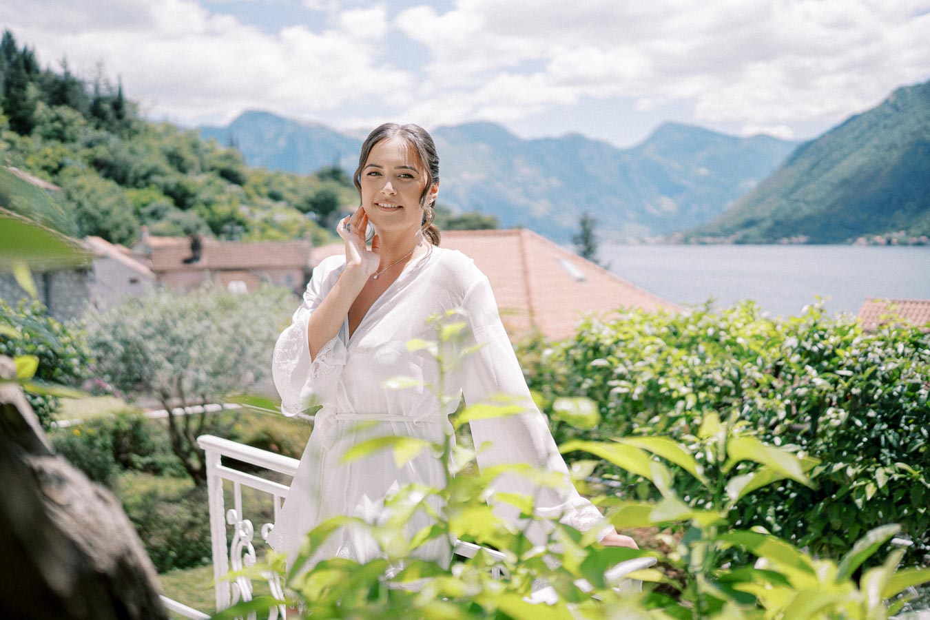 Woman in a white robe posing outdoors on a sunny day with a scenic mountain and lake view in the background, surrounded by lush greenery.