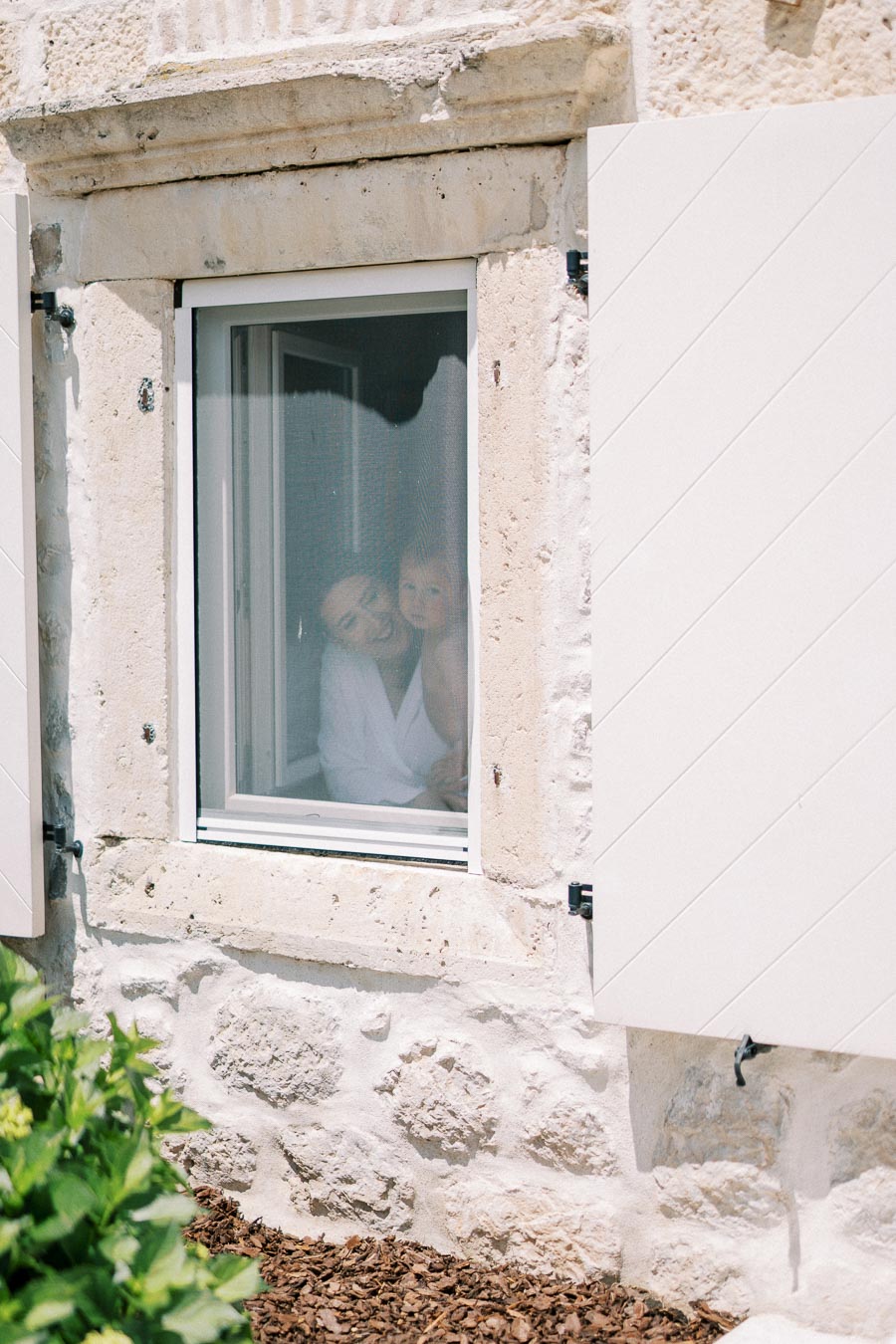 Mother and child smiling while peeking through a window of a rustic stone house with white shutters and greenery outside.