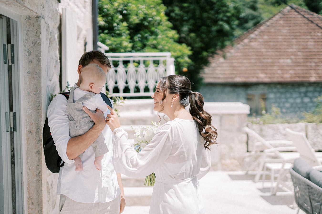 A woman in a white dress smiling at a man holding a baby, outdoor setting with stone architecture and greenery in the background.