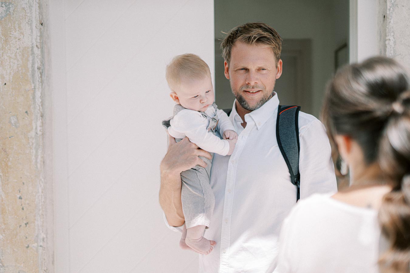 A man in a white shirt holding a baby at the entrance of a home, smiling at a woman in the foreground.