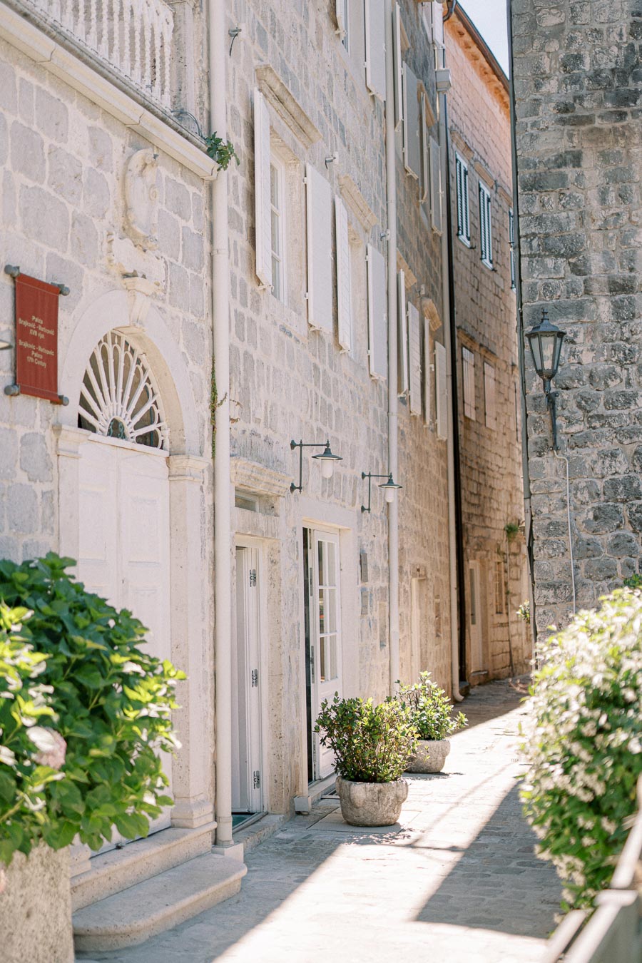 Charming narrow alleyway with historic stone buildings, adorned with potted plants and vintage lanterns, in a quaint European village on a sunny day.