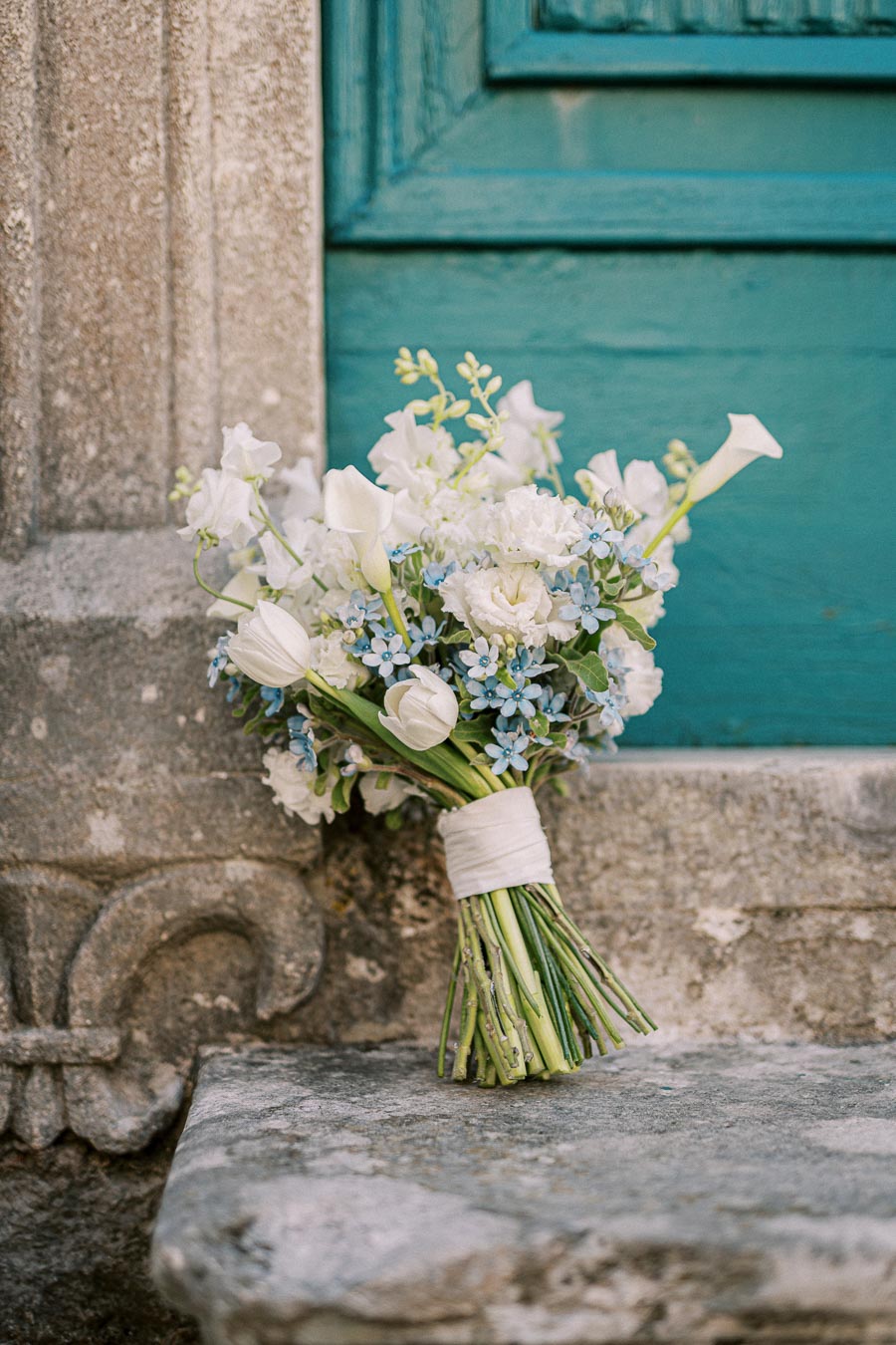 A rustic bouquet of white and blue flowers, including roses and lilies, rests against an aged stone wall beside a teal wooden door, creating a charming and vintage ambiance.