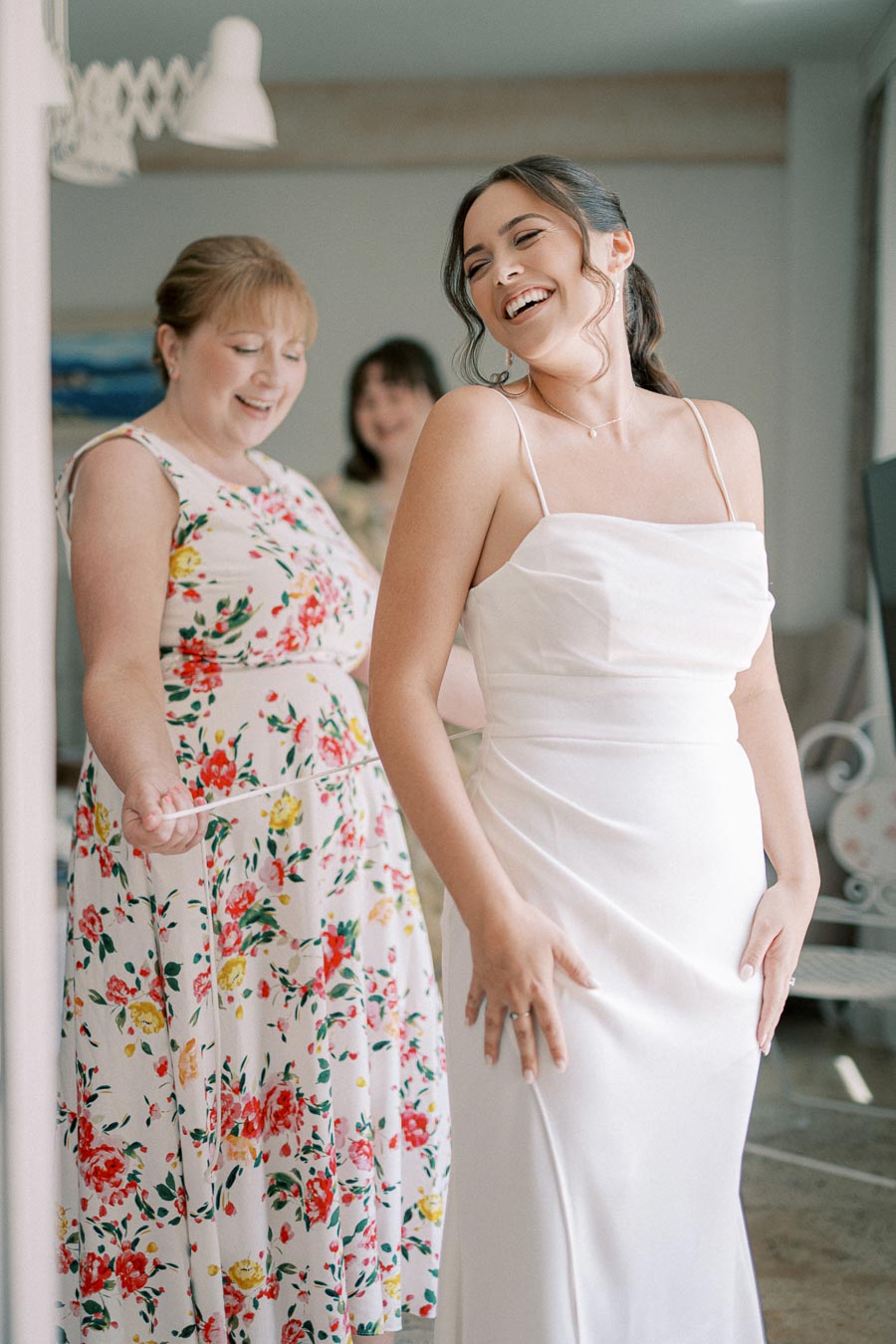 Bride in a white wedding dress smiling with bridesmaids wearing floral dresses in a bright room.