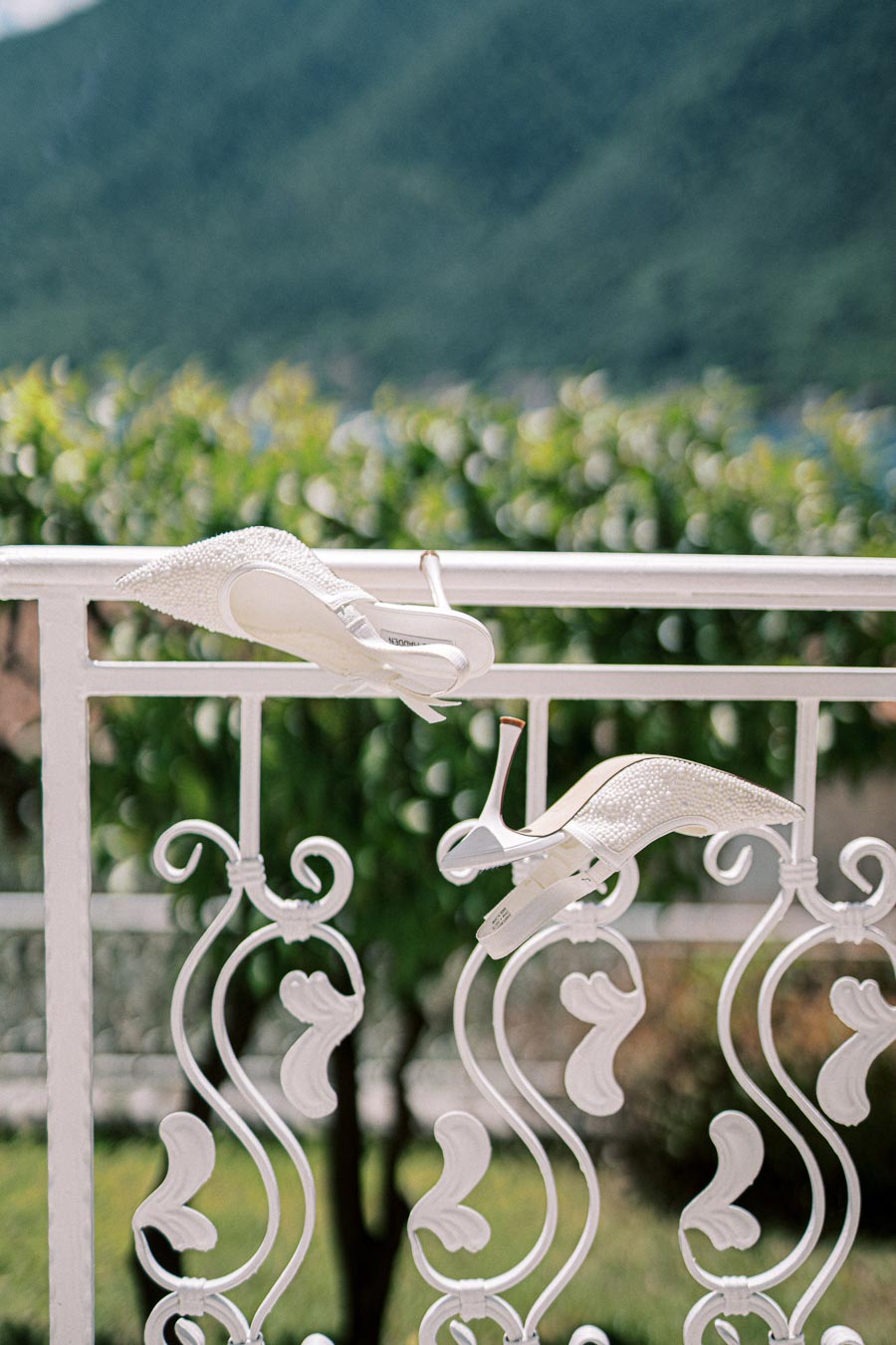 Pair of elegant white high heels with pearl embellishments resting on a decorative white wrought iron railing, with a scenic green mountain landscape in the background.