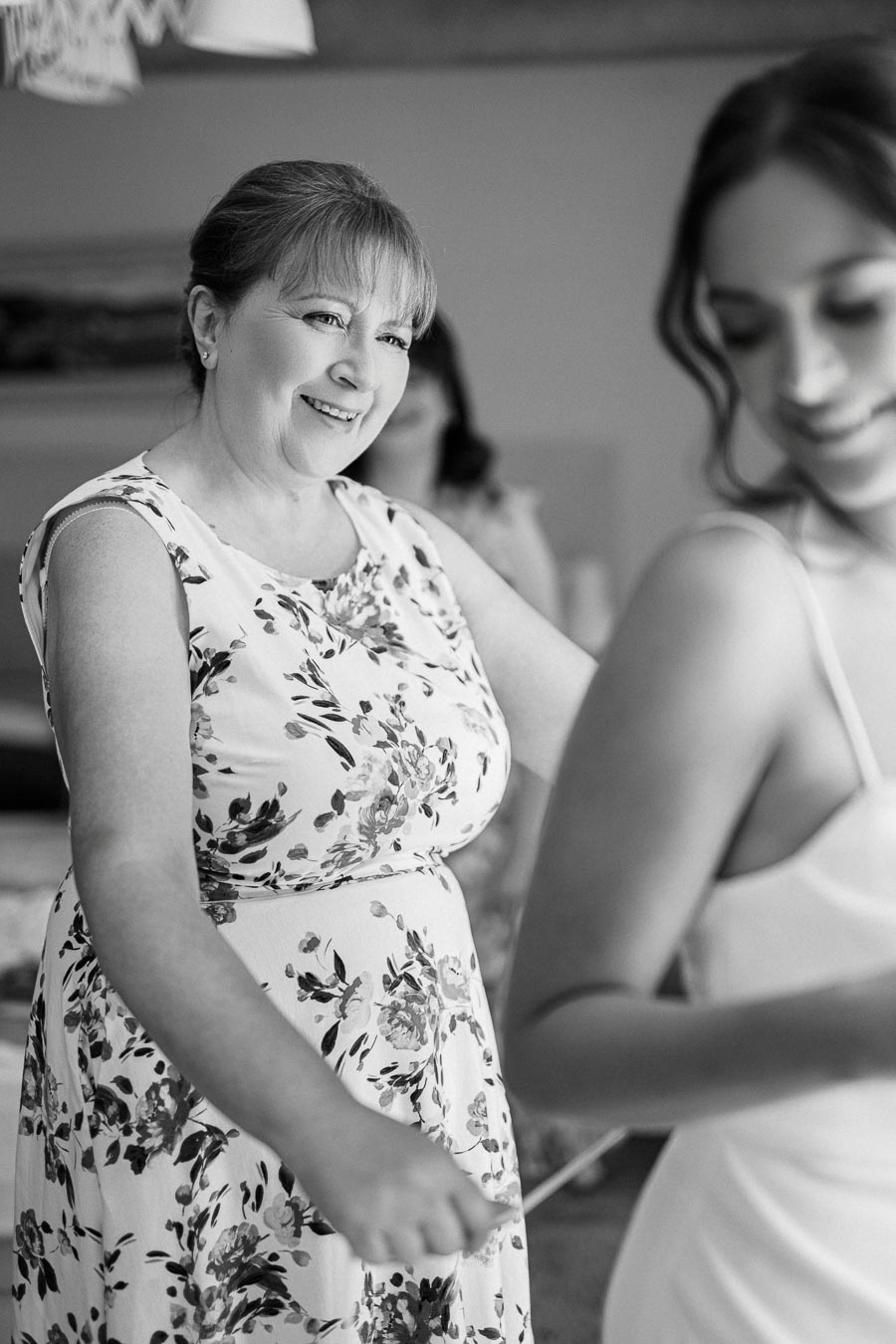 Joyful woman in floral dress helping bride with wedding preparation, captured in a candid black and white moment.
