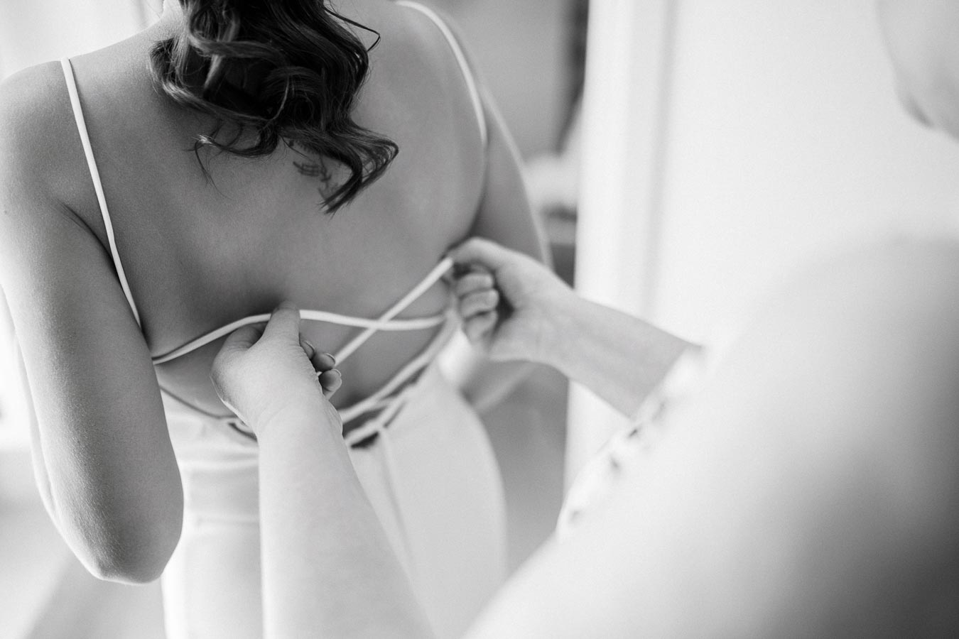 Black and white close-up of a bride having her wedding dress adjusted from the back, highlighting intricate lace-up details and elegant straps.