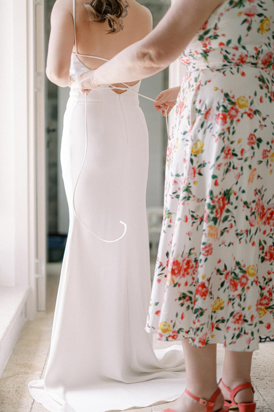 A woman in a floral dress helps another woman lace up the back of a white wedding gown, preparing for a wedding ceremony in a softly lit room.