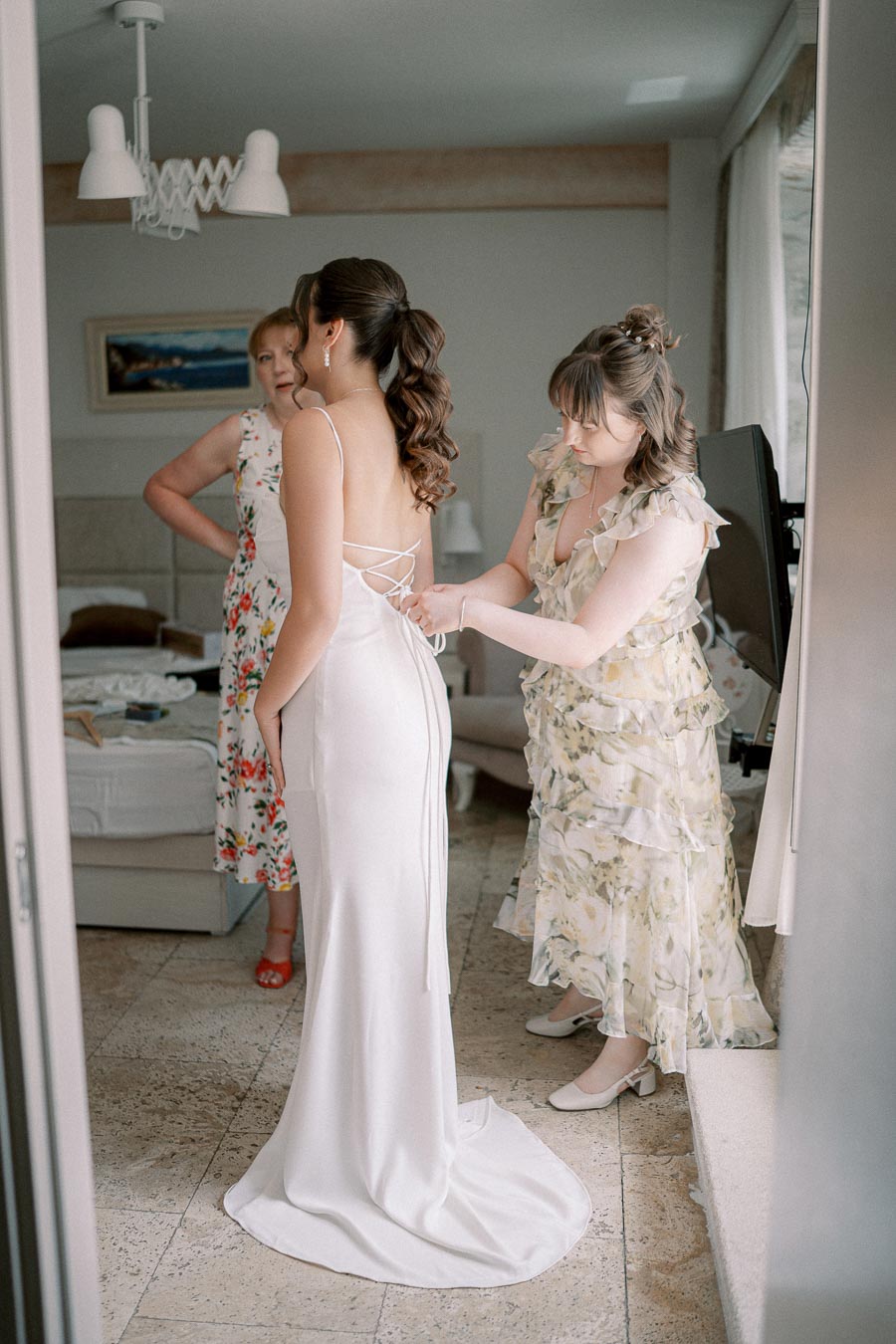 Bridesmaids help bride in elegant white wedding dress prepare for ceremony in a bright room.