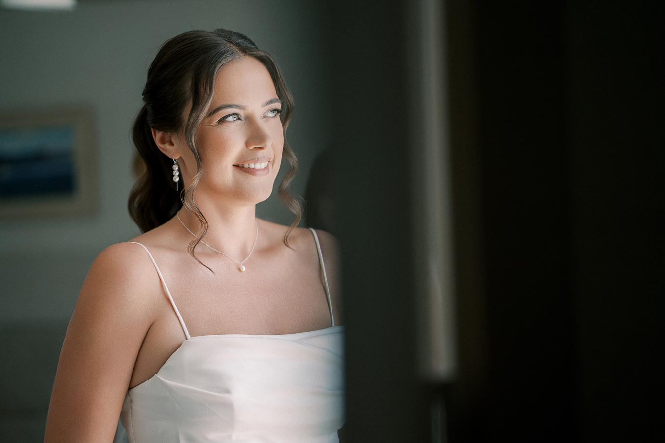 Smiling bride in elegant white dress with pearl earrings gazing towards a window.