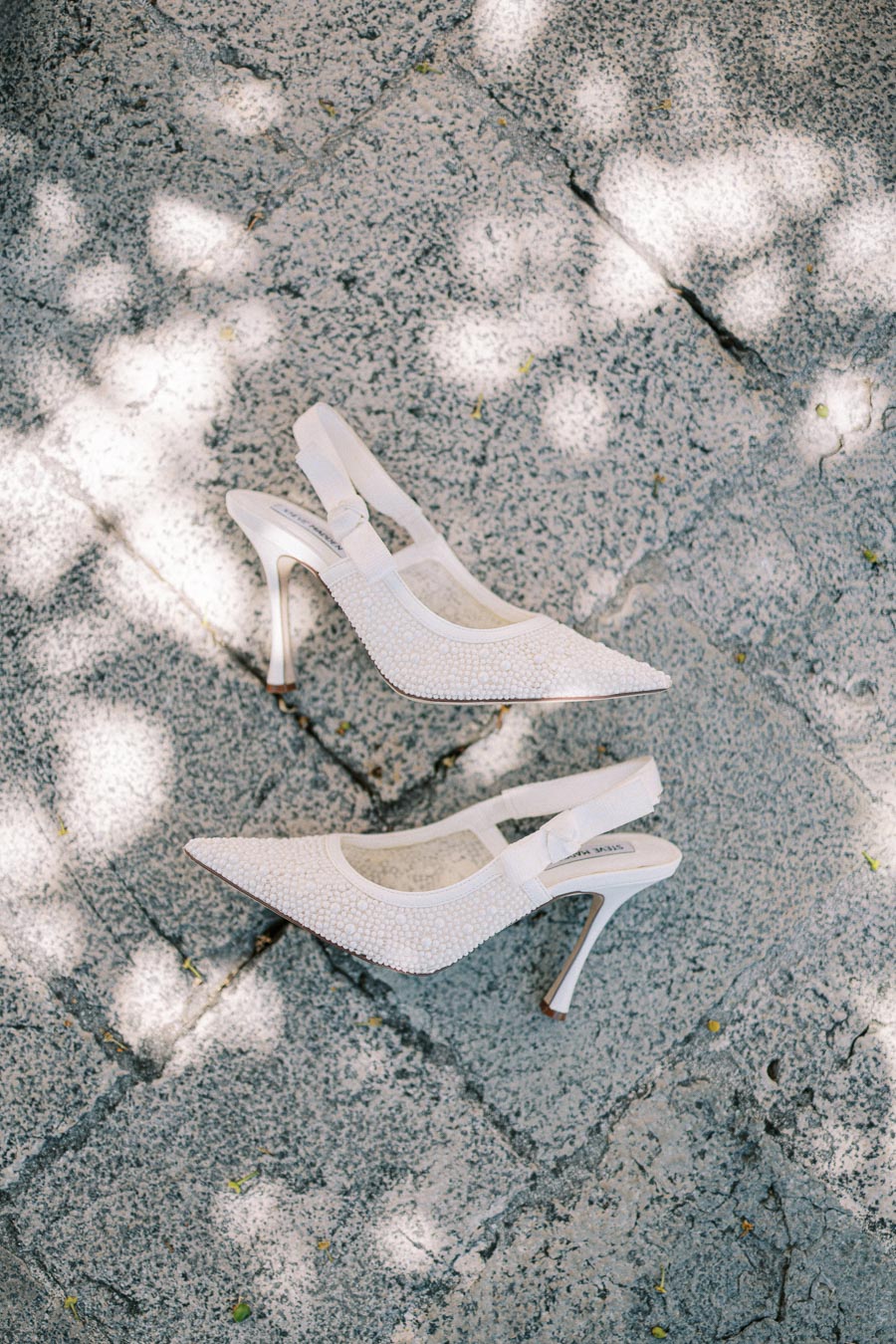 Elegant white high-heeled shoes with intricate beadwork, placed on a textured stone surface dappled with sunlight, perfect for a stylish bride or formal event.
