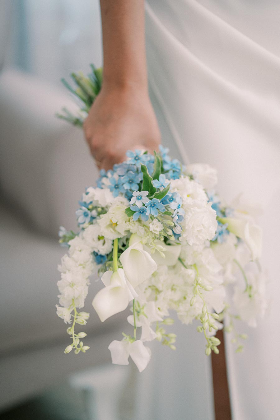 Bridal hand holding a bouquet of blue forget-me-nots, white carnations, and calla lilies against a soft, blurred background.
