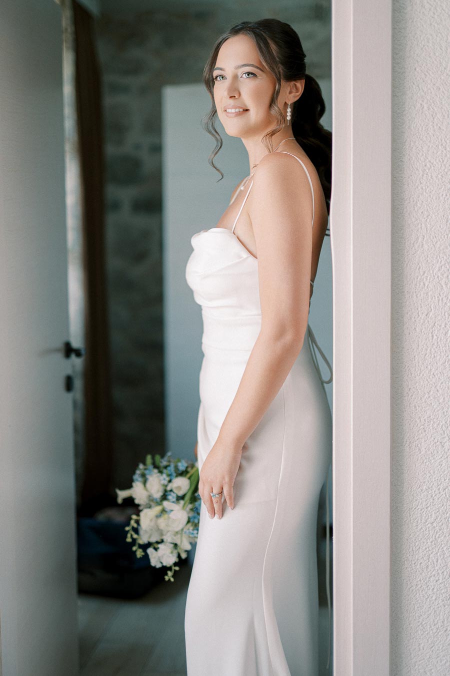 A bride in an elegant white dress holding a bouquet of white and blue flowers, standing indoors by a doorway with soft natural light.