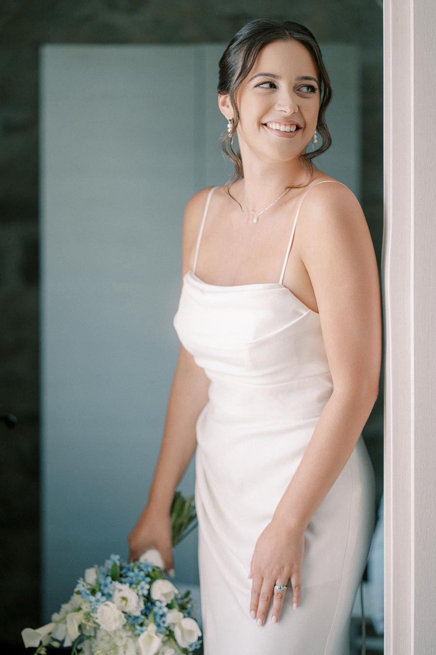 A smiling bride in a white wedding dress holding a bouquet of white and blue flowers, standing near a doorway.