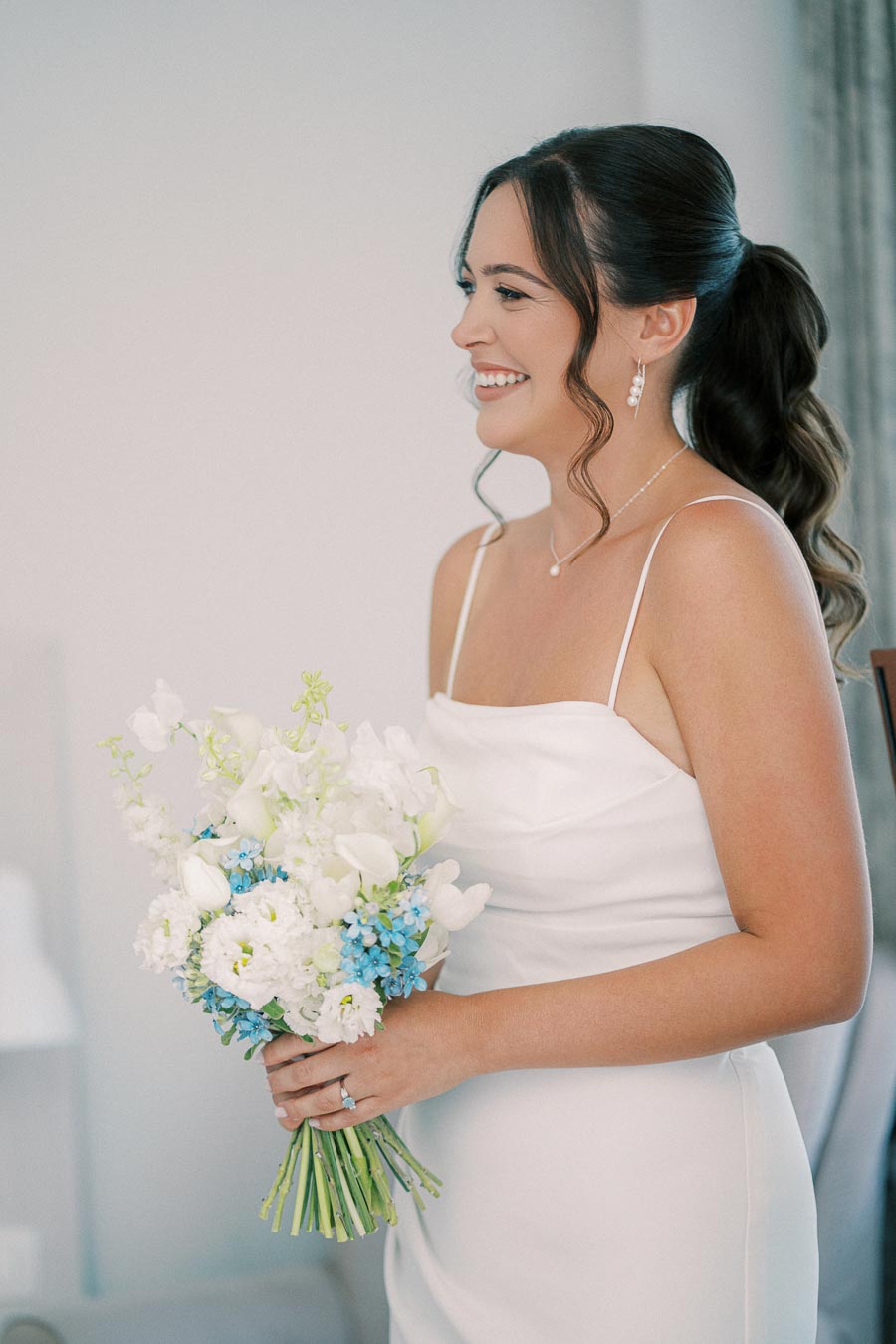 A bride in a white wedding dress smiling while holding a bouquet of white and blue flowers, showing elegant bridal jewelry and a modern hairstyle.