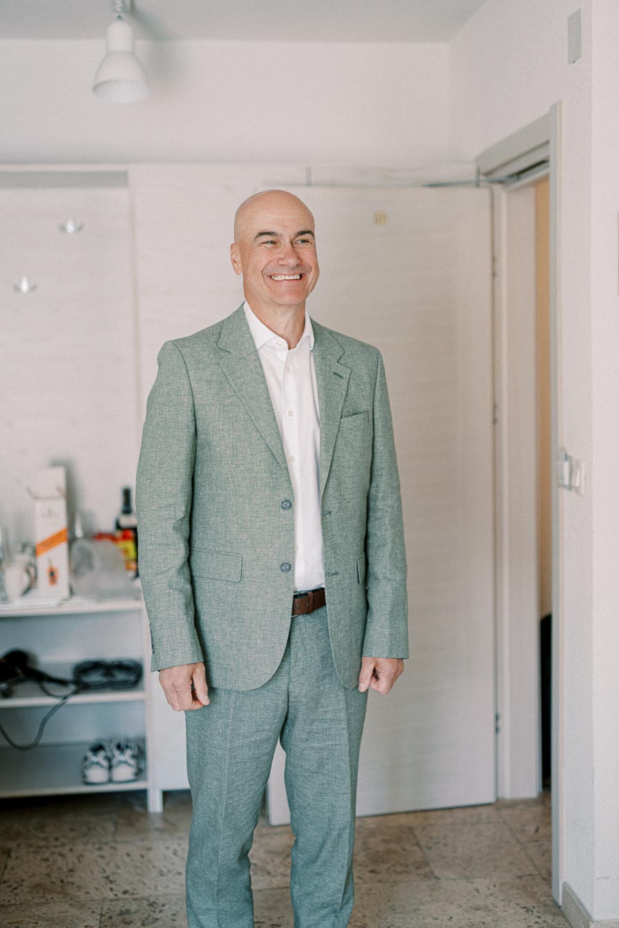 A man in a green suit smiling confidently, standing in a modern room with shelves and wooden flooring.