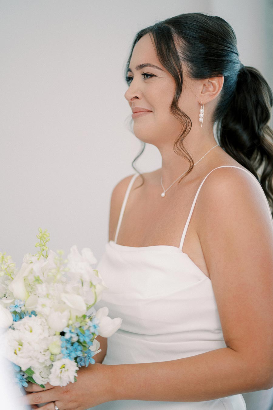 A bride in a white dress holding a bouquet of white and blue flowers, smiling softly with elegant pearl earrings and a delicate necklace.