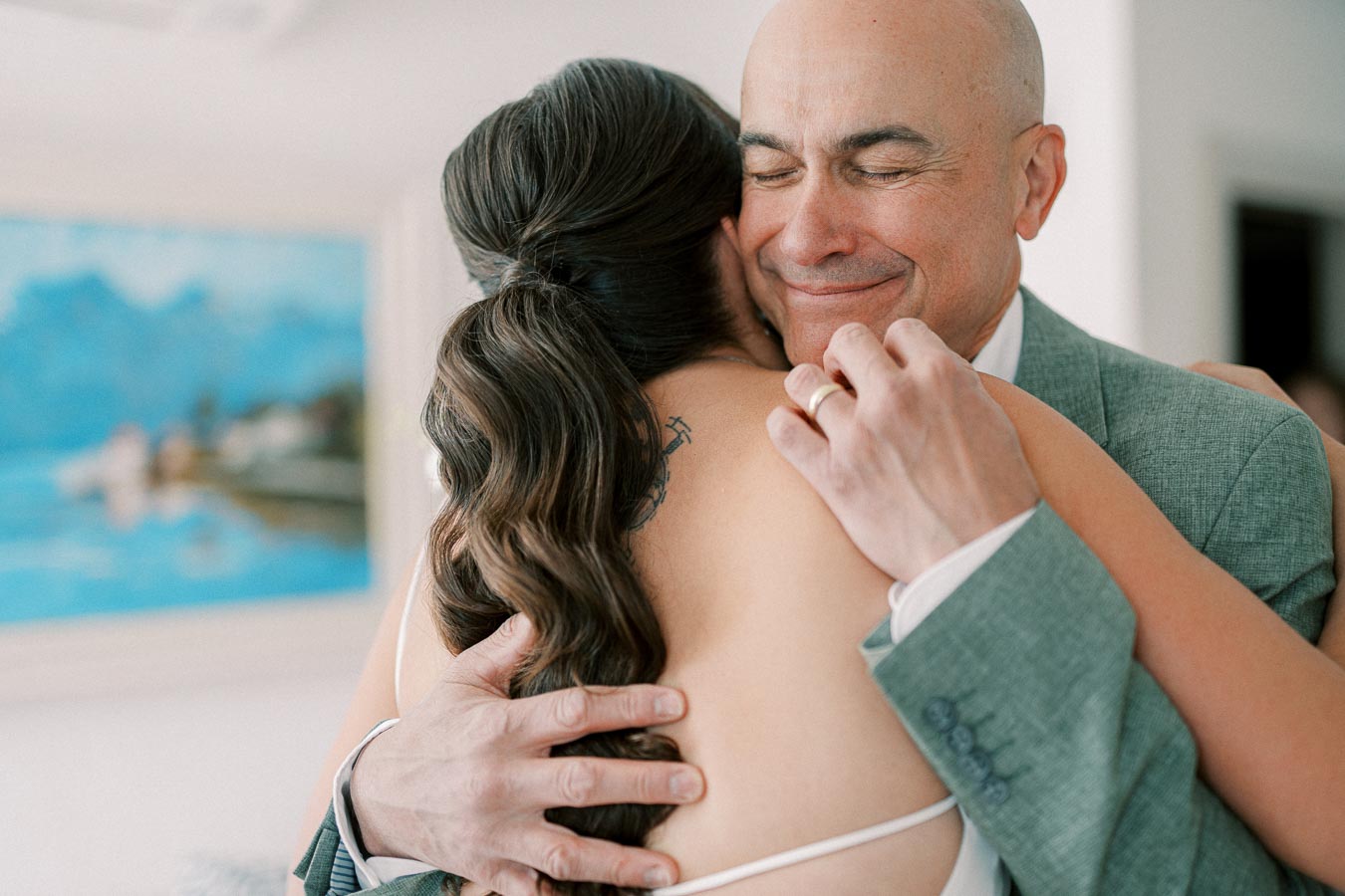Father and daughter sharing an emotional hug on a wedding day; the father is wearing a gray suit while the bride with long brown hair wears a white dress, capturing a heartfelt moment of love and joy.
