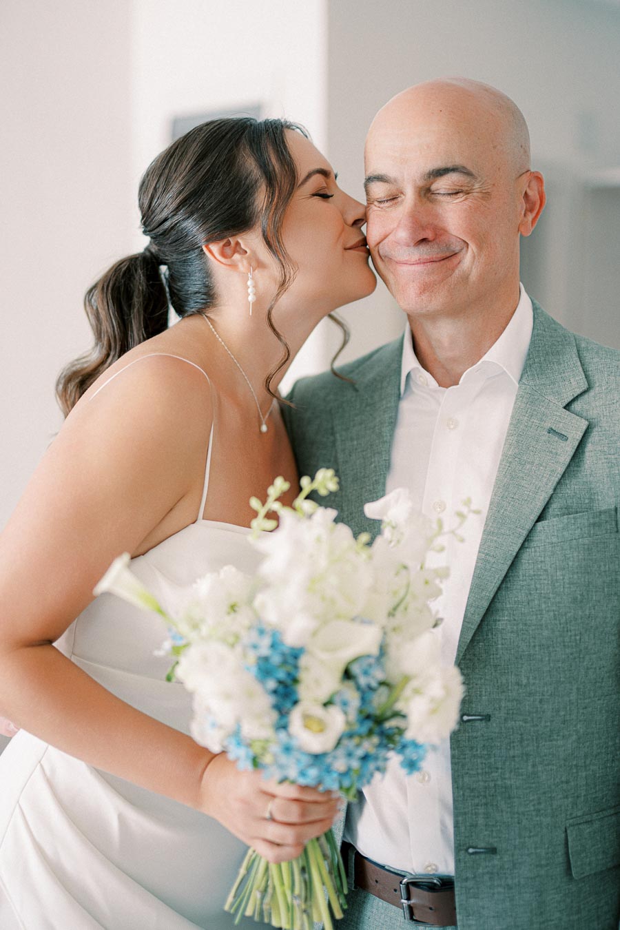 A bride in a white dress kisses a smiling man in a light gray suit, holding a bouquet of blue and white flowers.