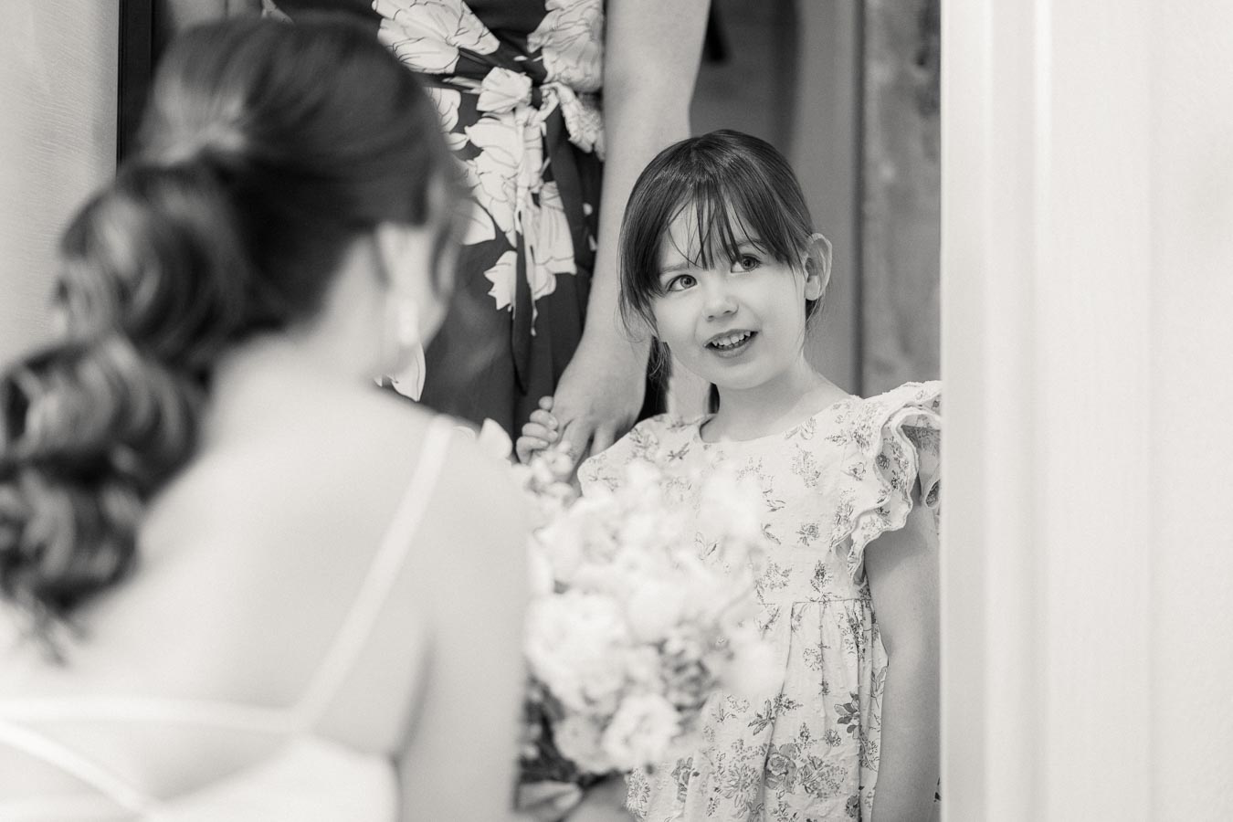 Black and white photo of a young girl in a floral dress smiling at a woman holding a bouquet, capturing a tender moment before a wedding ceremony