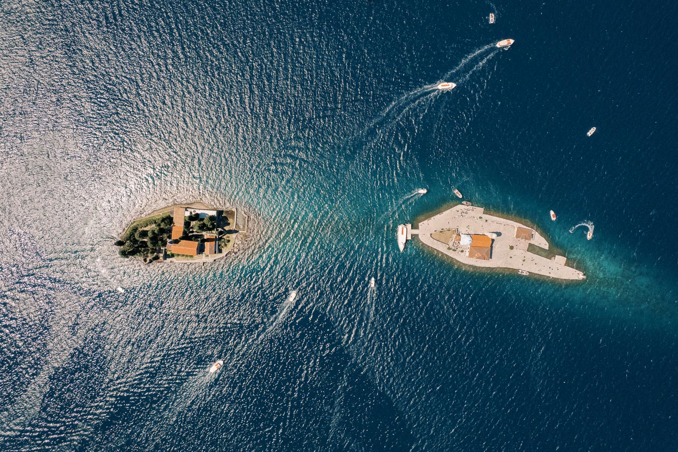 Aerial view of two small islands in the Adriatic Sea, featuring historic buildings with terracotta roofs surrounded by deep blue water and small boats navigating between them.