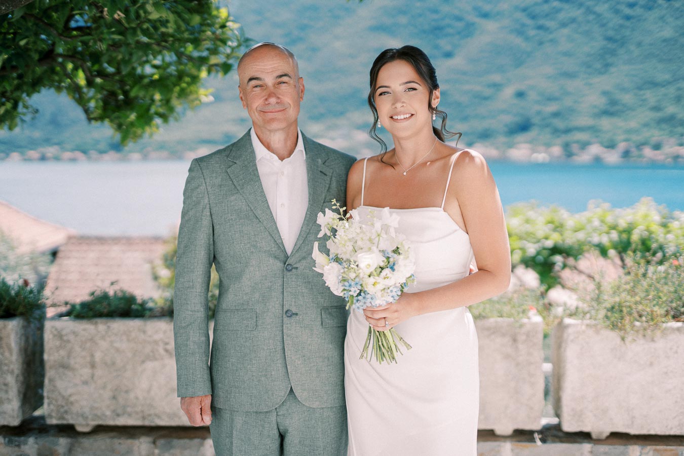 Father and bride at outdoor wedding ceremony, smiling and standing by a scenic waterfront view; she is holding a bouquet of white and blue flowers.
