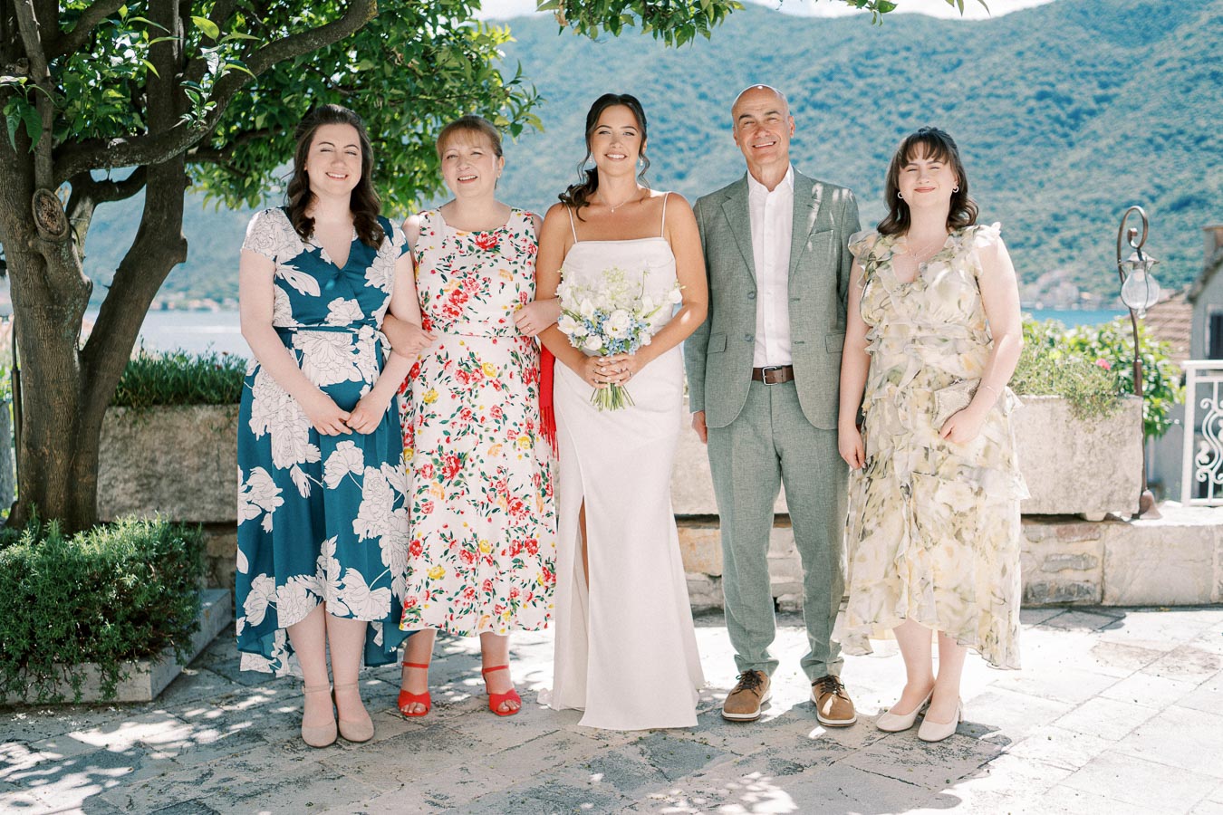 A group of five people posing together outdoors at a wedding, with the bride in a white gown holding a bouquet and surrounded by family members in colorful dresses and a suit, set against a scenic mountain and water background.