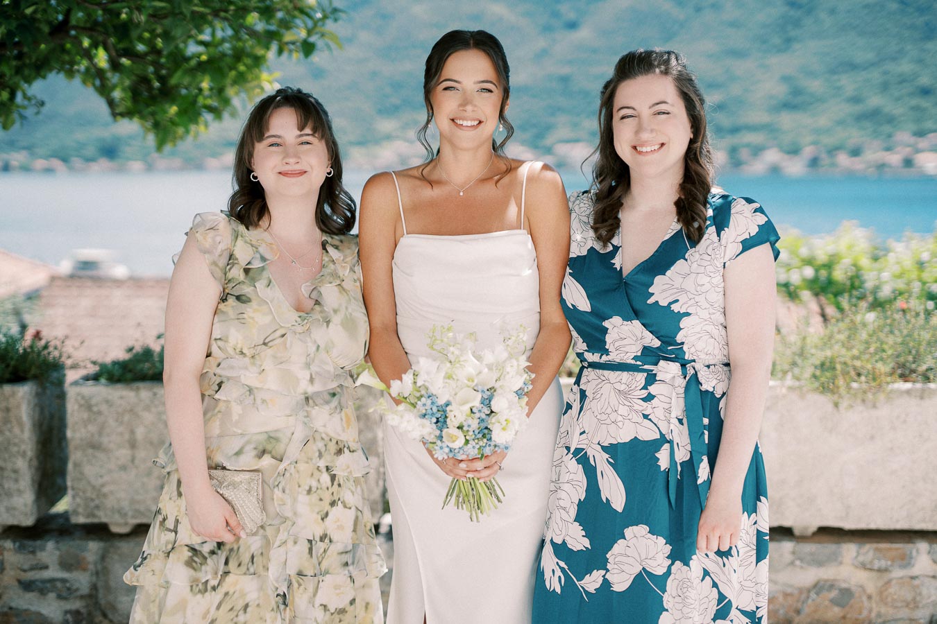 Three women standing together outdoors, one in a white dress holding a bouquet, surrounded by greenery and a scenic lake view.