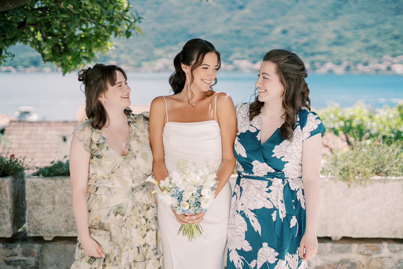 Three women smiling outdoors on a sunny day. The woman in the center is wearing a white dress and holding a bouquet of white and blue flowers, flanked by two women in floral dresses. Scenic background of a lake and mountains. Perfect for wedding or celebration themes.