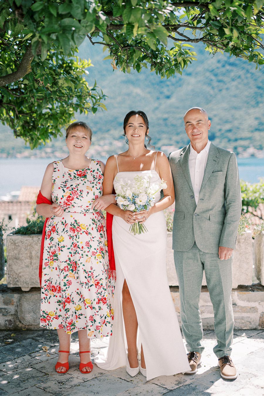 A bride in a white gown poses with two people under a leafy tree, overlooking a scenic waterfront view. The woman on the left wears a floral dress with red accents, and the man on the right is in a light gray suit. The background features mountains and a tranquil body of water.