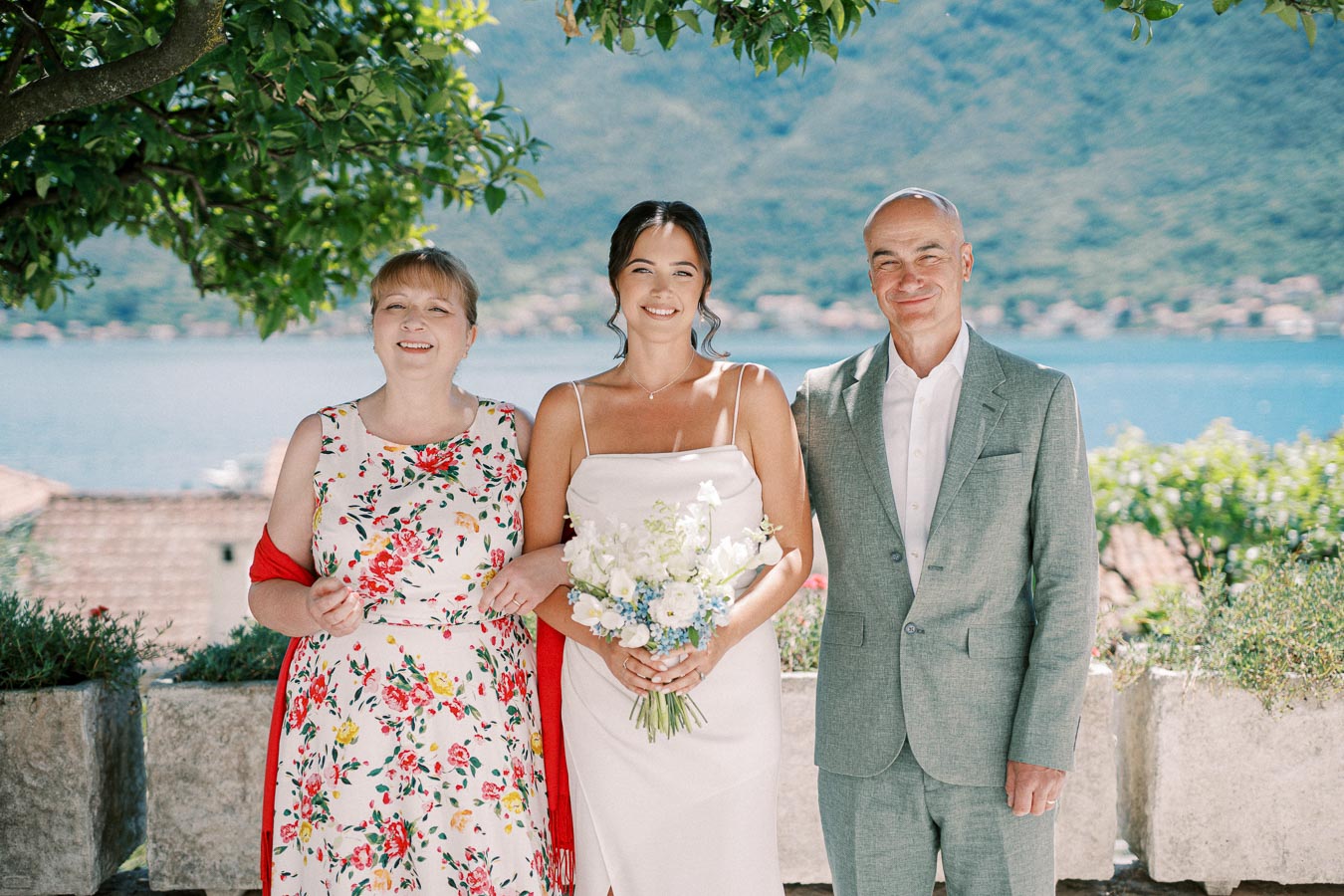 A joyful bride in a white dress holding a bouquet stands between her smiling parents, with lush greenery and a picturesque lake in the background.