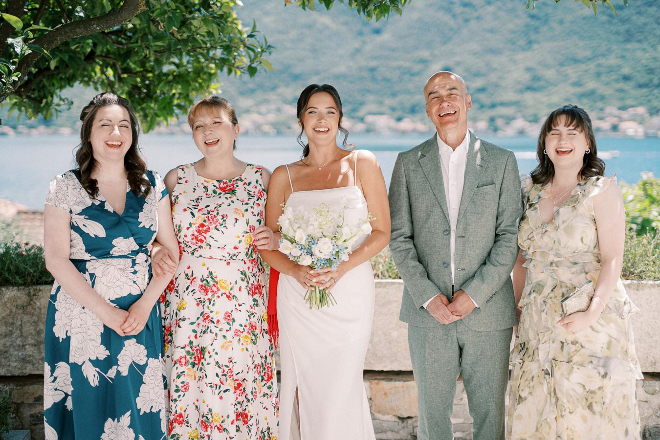 A group of five people, including a bride holding a bouquet, standing outdoors by a scenic waterfront with mountains in the background, dressed in formal attire.