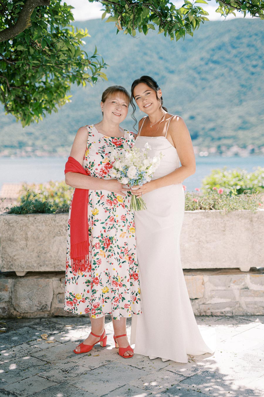 Two women smiling at a wedding, one in a floral dress and red shawl, and the other in a white bridal gown holding a bouquet, with a scenic lake and mountain background.