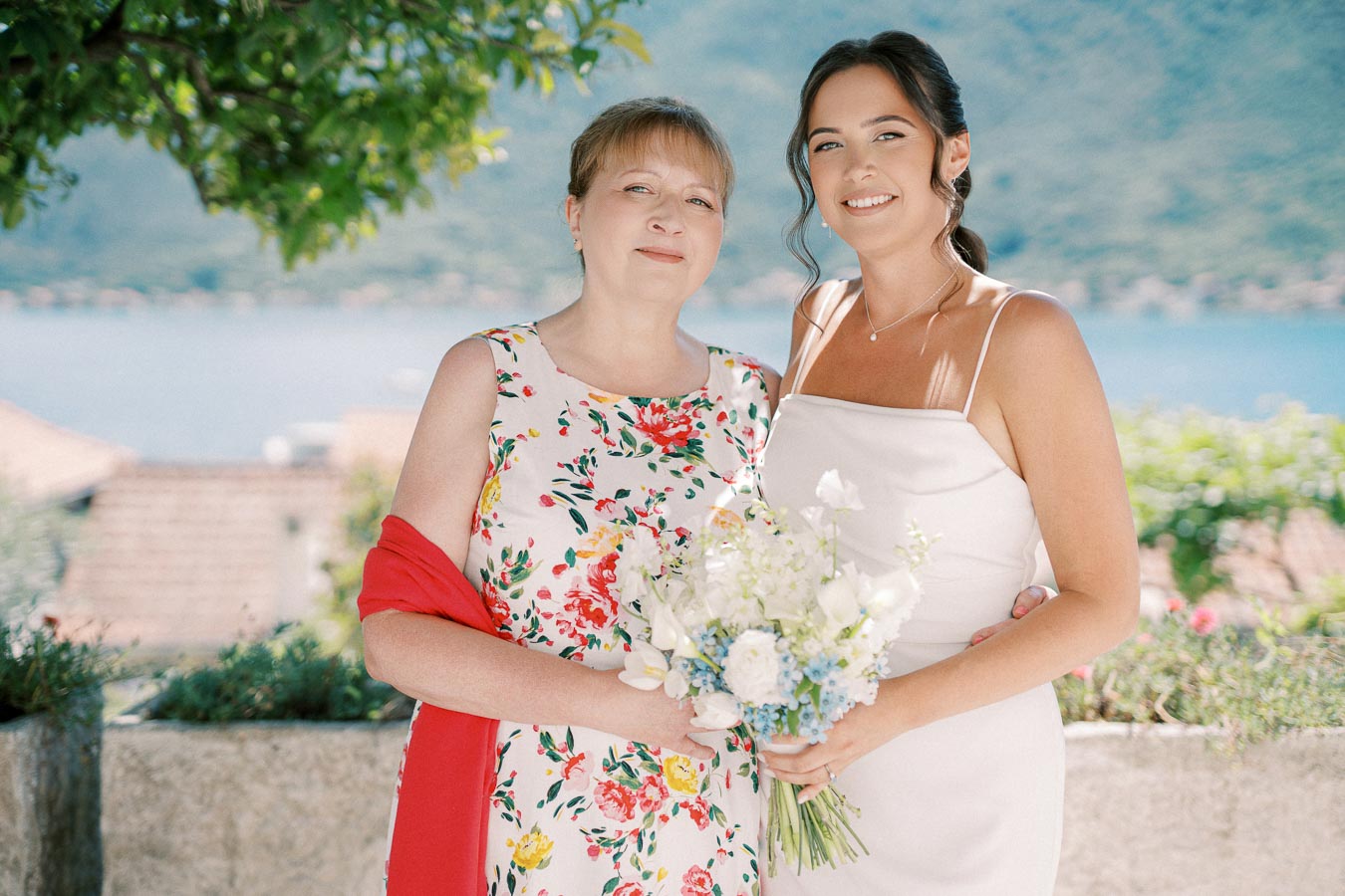 Smiling woman in white dress holding bouquet stands beside another woman in floral dress with red shawl, with scenic lakeside background.