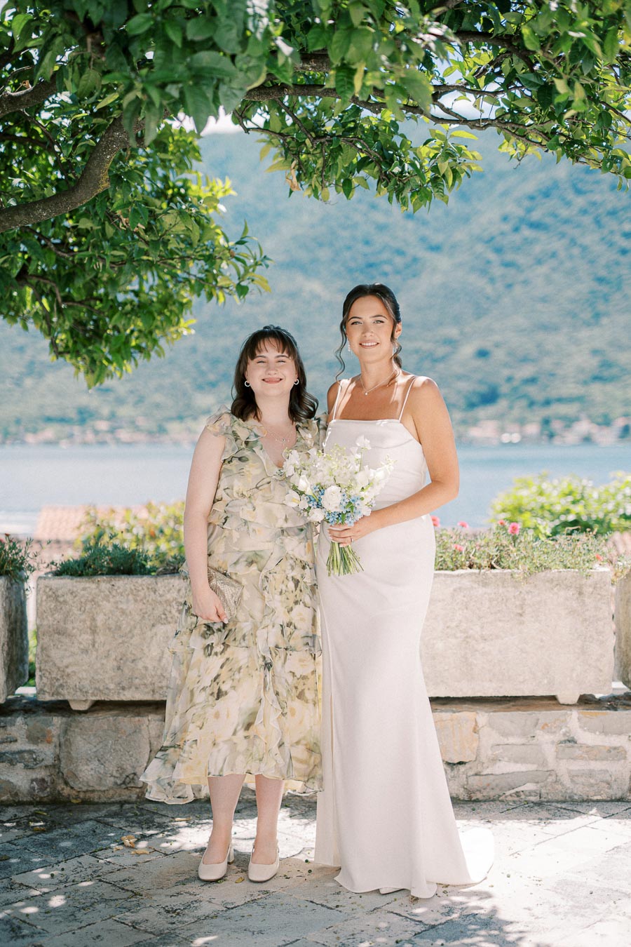Two women standing outdoors under a tree, with a scenic background of a lake and greenery. One wears a floral dress and the other a white gown, holding a bouquet of flowers.