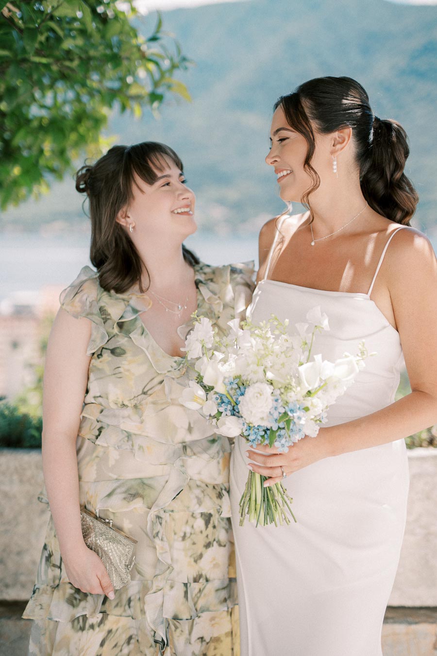 Two women smiling at each other outdoors, one in a white dress holding a bouquet of white and blue flowers, with a scenic mountain background.