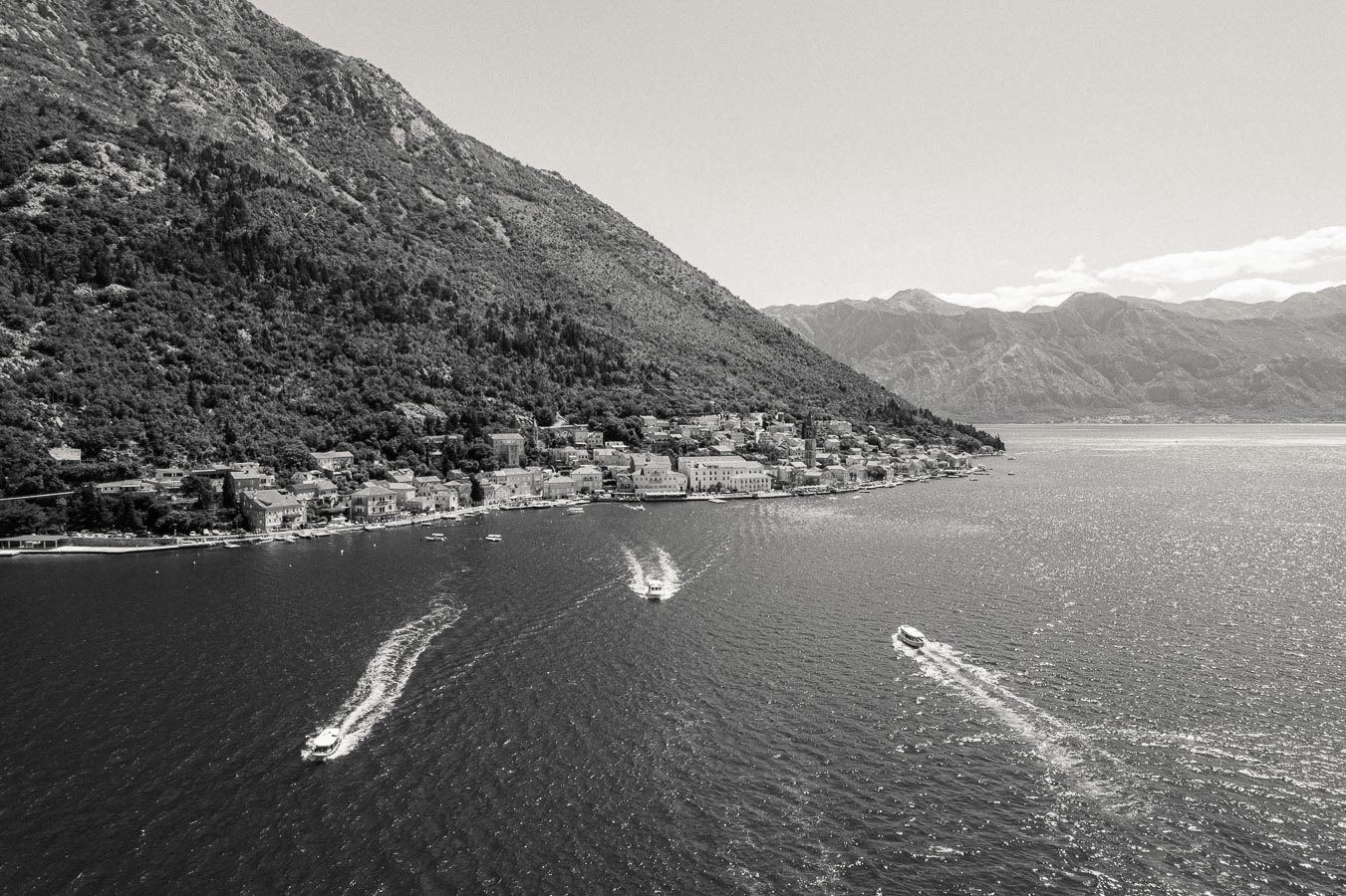 Black and white aerial view of a coastal town nestled at the base of a lush, mountainous landscape, with boats leaving trails on the shimmering water, capturing the tranquility and scenic beauty of the seaside community.