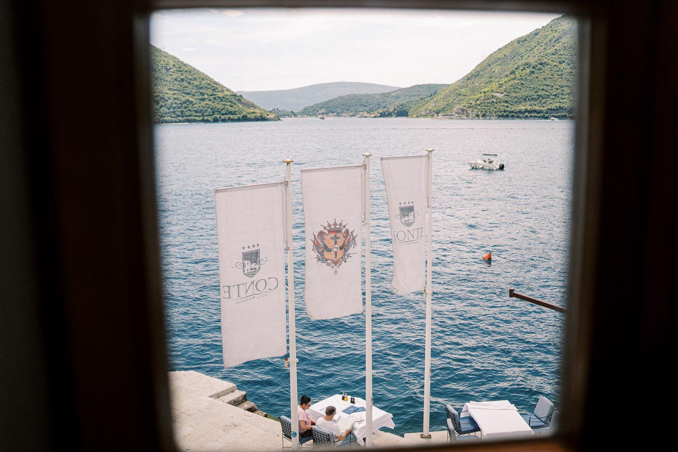 View through a window of a serene waterfront dining area with three flags. Two people sitting at a table near the water, surrounded by scenic hills and a small boat in the distance.