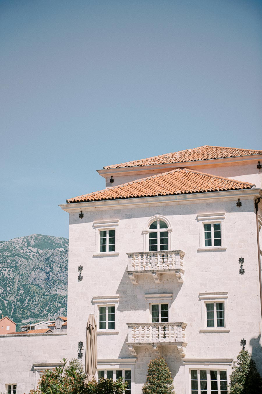 Elegant stone building with balconies and terracotta roof against a backdrop of mountains and clear blue sky.