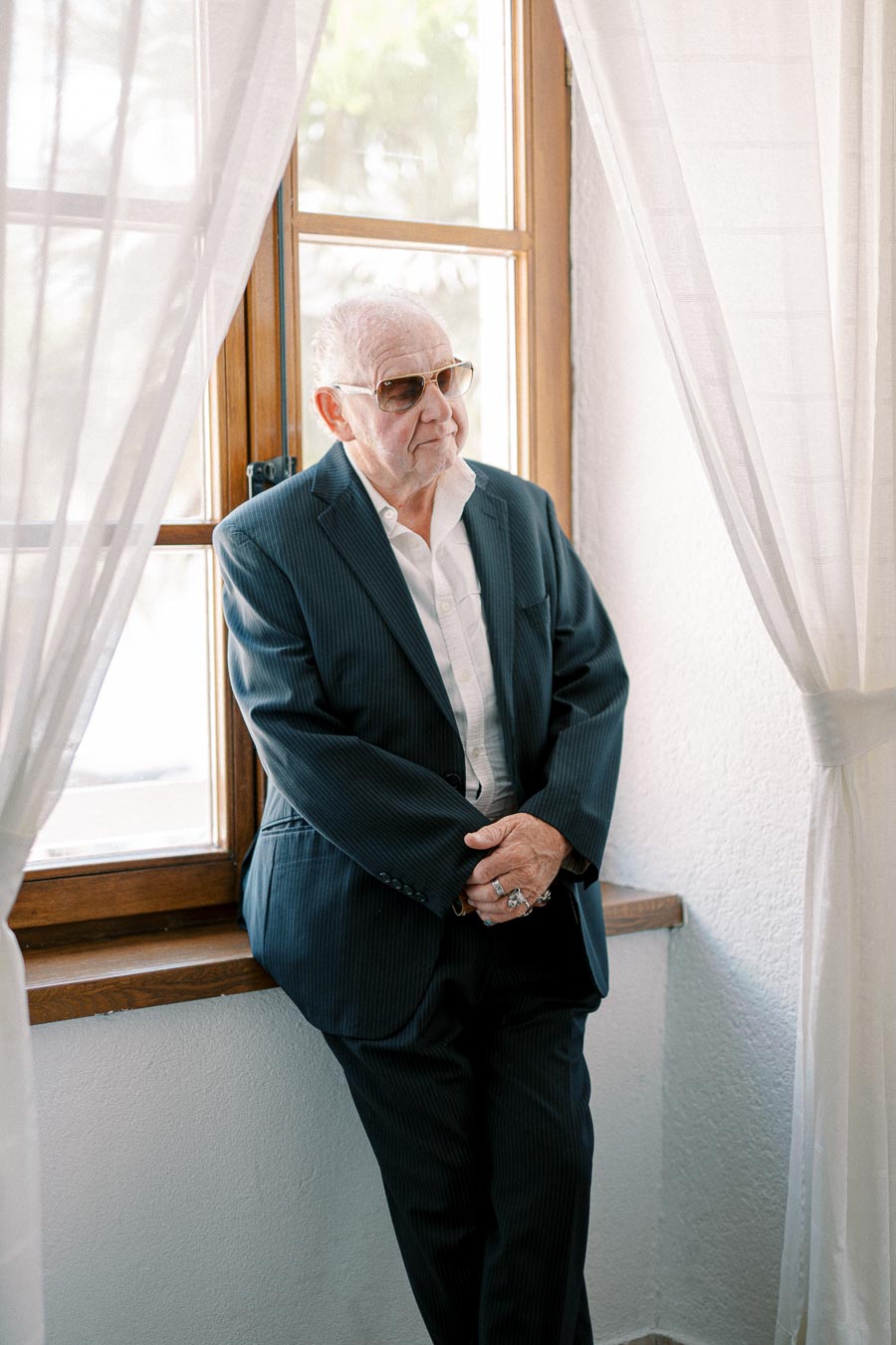 Elderly man in a suit with sunglasses standing by a window with sheer curtains, exuding a sense of style and confidence.