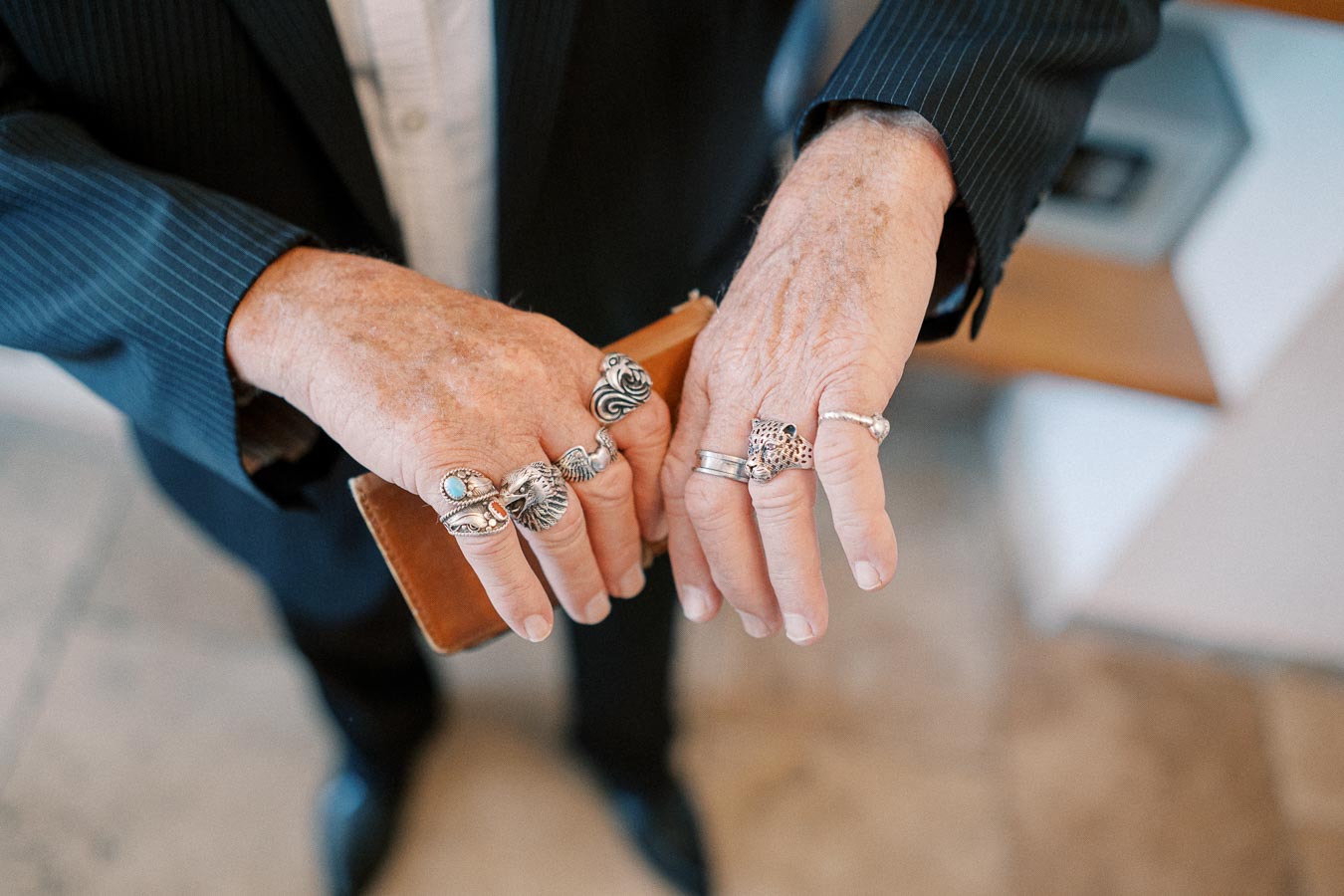 Close-up of a person's hands adorned with multiple unique silver rings, each featuring intricate designs and stones, while holding a wallet and wearing a pinstripe suit.